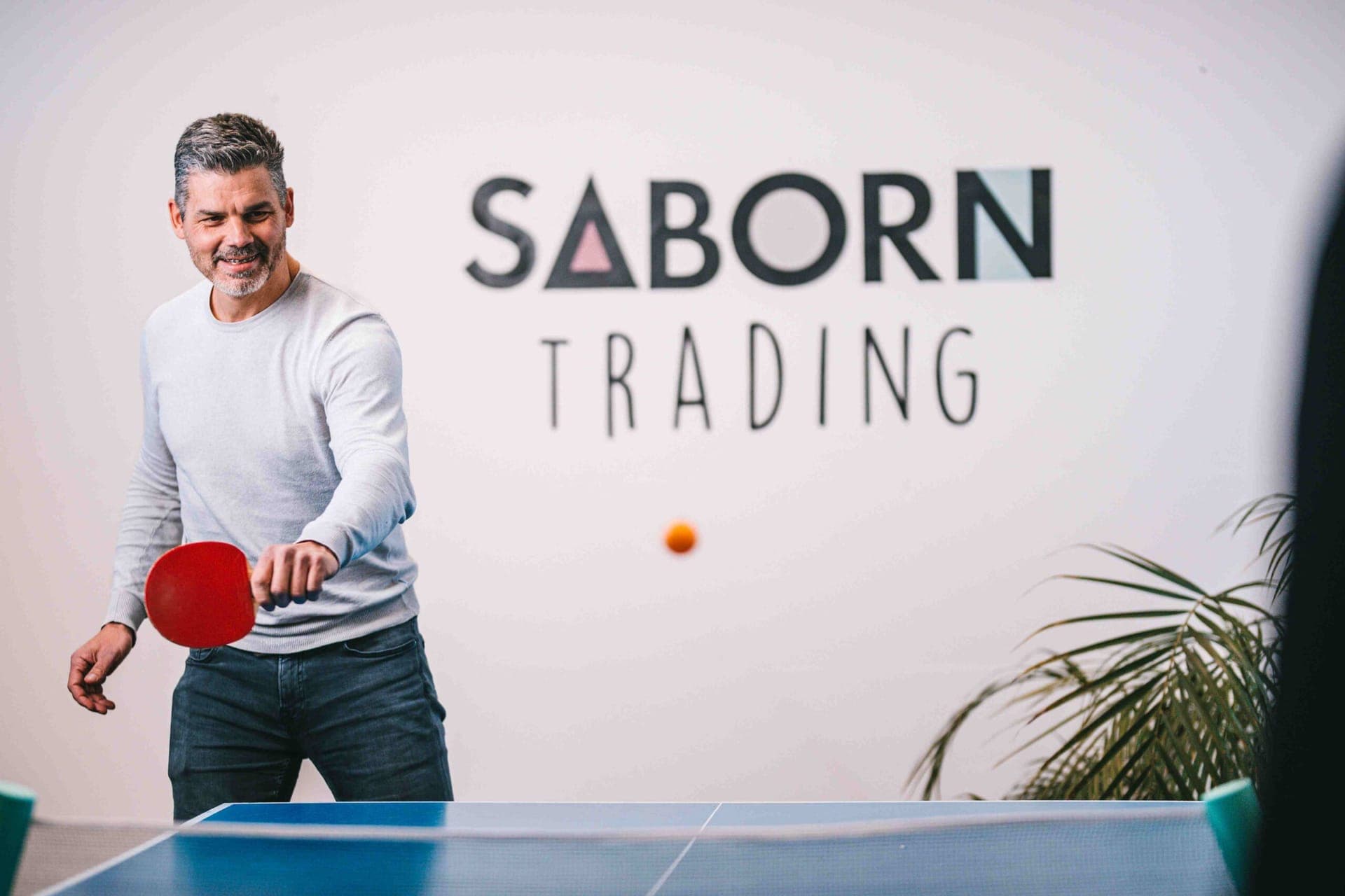 Collega speelt tafeltennis voor het Saborn Trading-logo op de kantoormuur